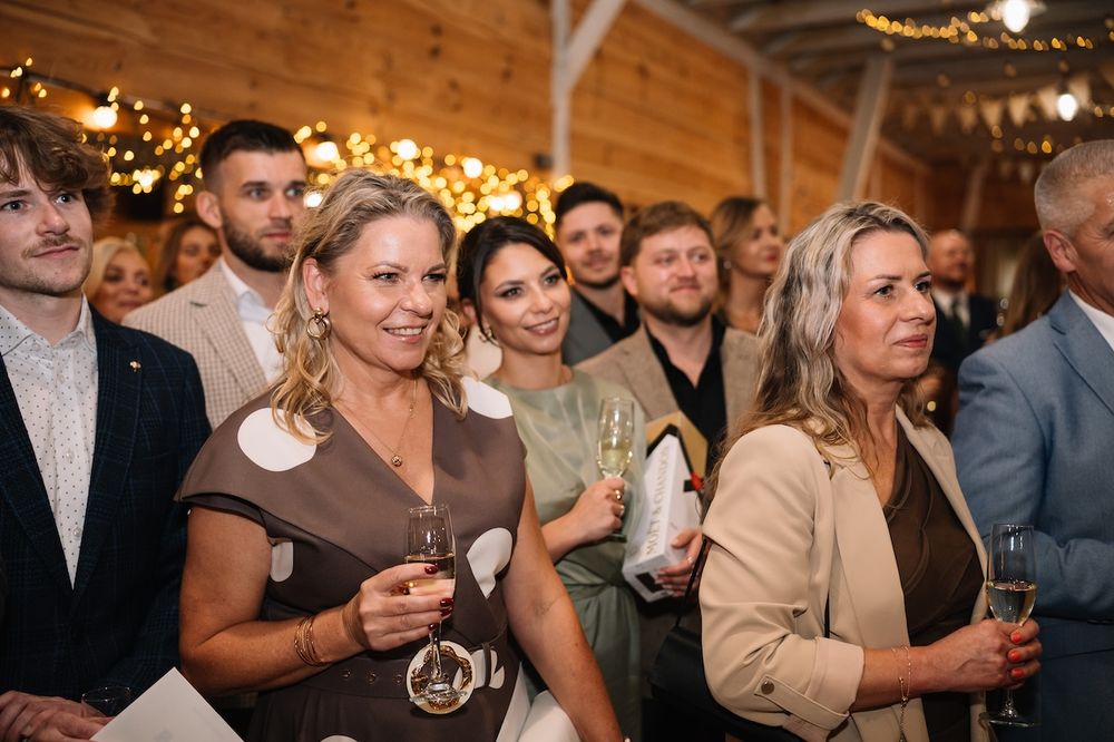 Wedding guests smile and hold champagne glasses during reception celebration in rustic decorated venue.