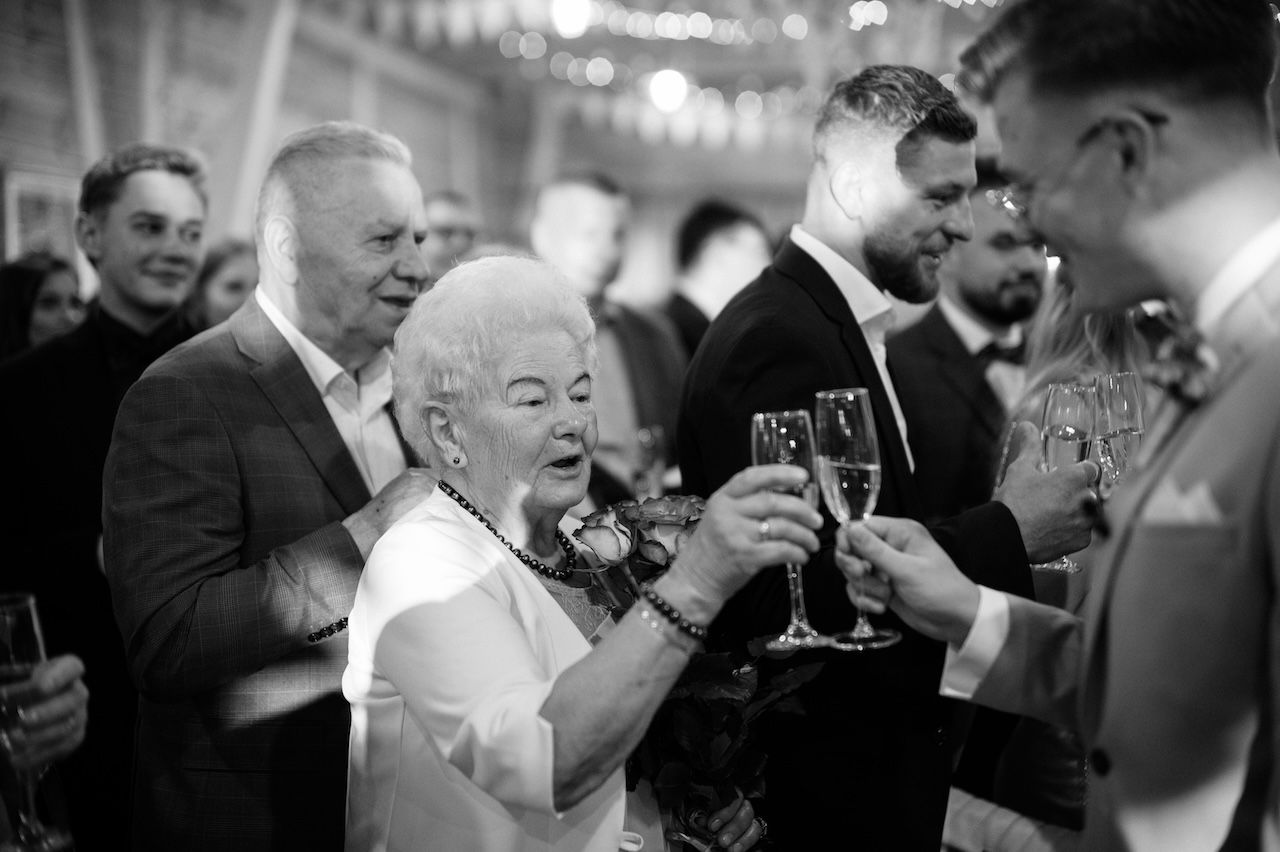 Black and white photo of elderly woman toasting champagne with wedding guests at reception.