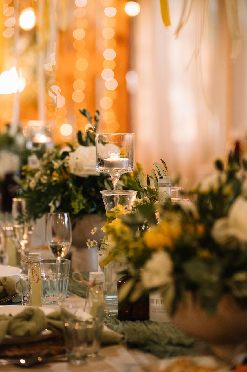 Elegant wedding table centerpiece with white and yellow flowers, candles, and warm bokeh lights.