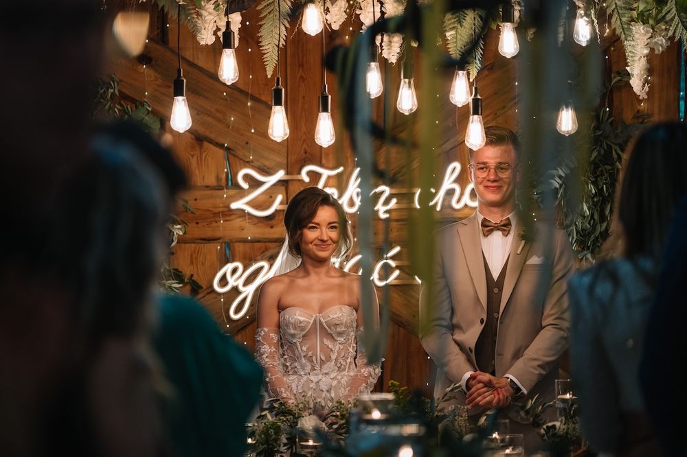 Bride and groom seated at wedding reception table under warm Edison bulb lights and neon sign.