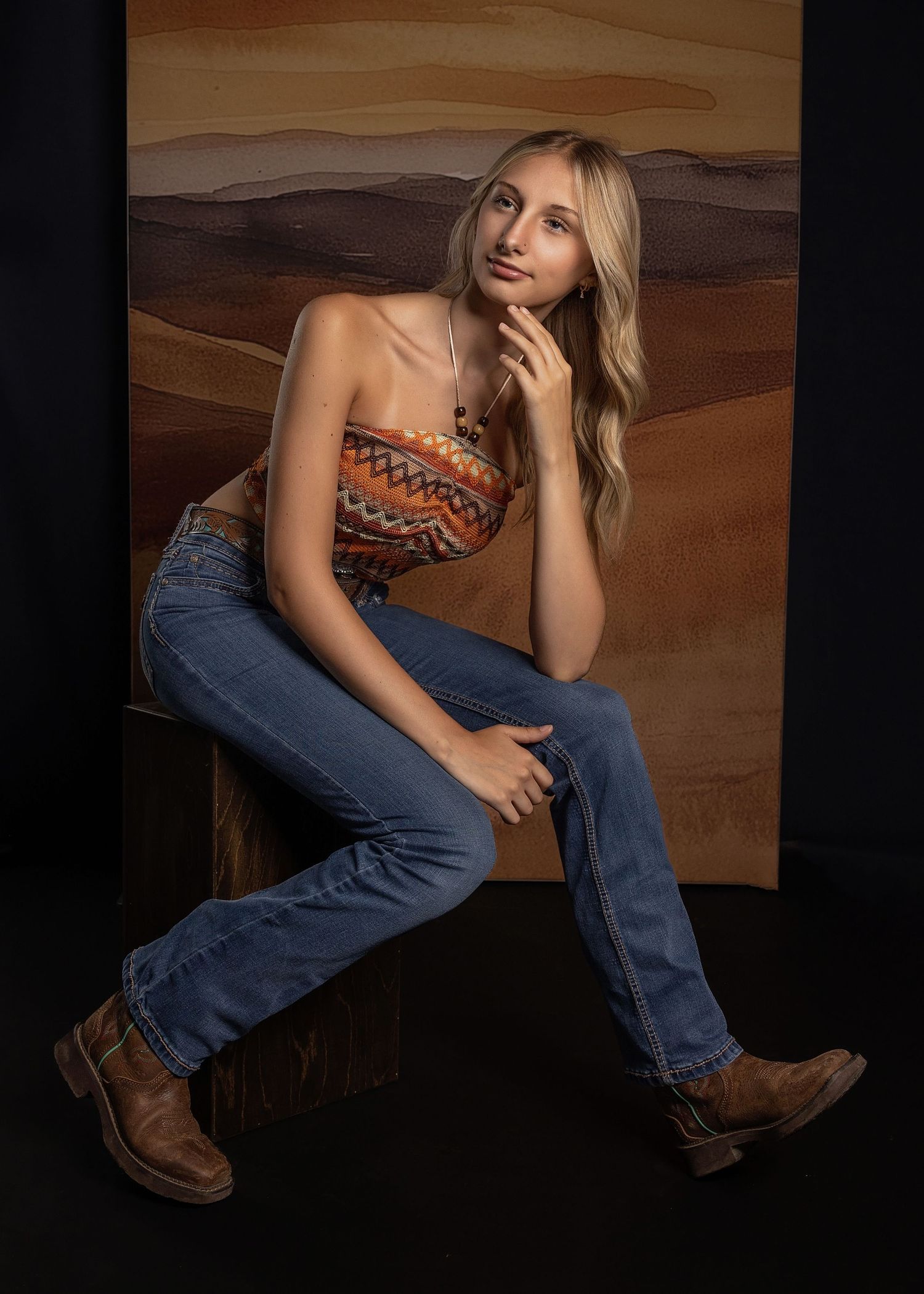 Fashion portrait against brown backdrop wearing patterned crop top, blue jeans and brown boots in casual pose.