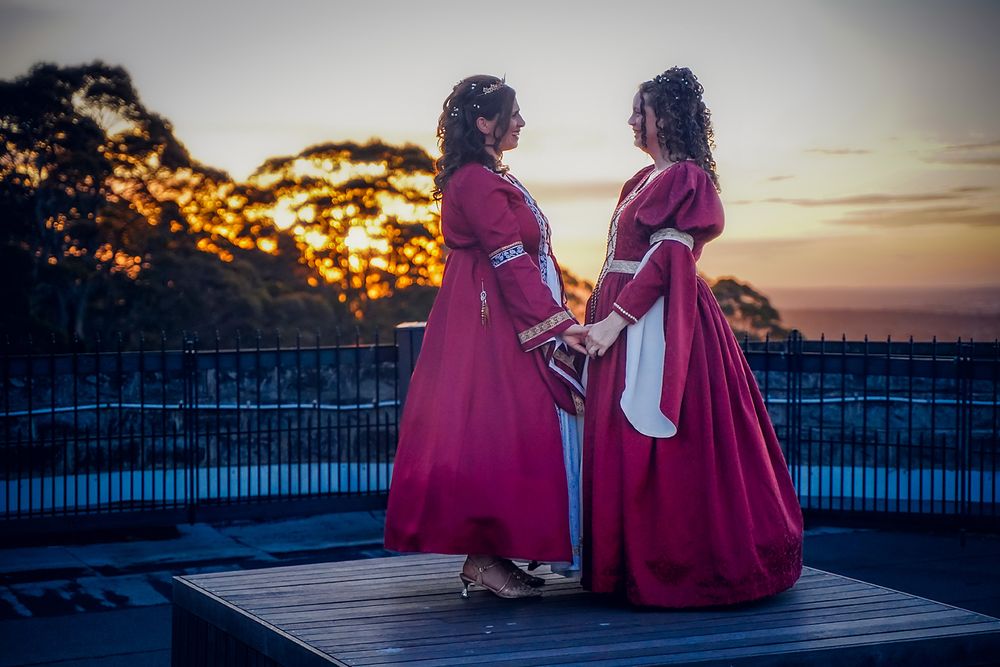 Two figures in pink dresses sit together on a deck at sunset creating a romantic silhouette against the sky.