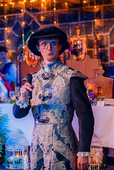A person in period costume wearing a black hat and ornate white lace collar dances at an elegant evening event.