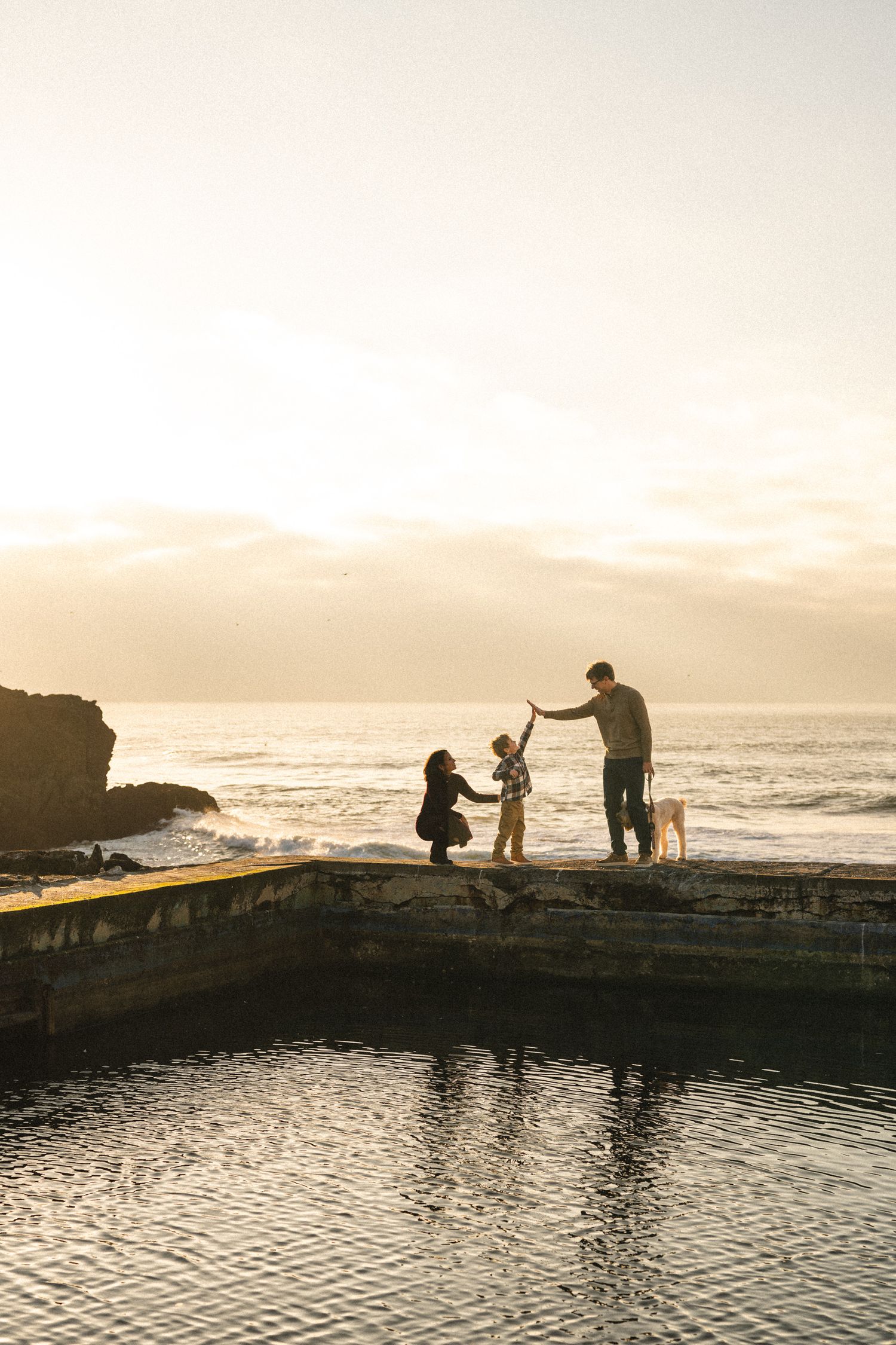 Silhouettes of people fishing at sunset by a coastal tide pool with mountains in the background.