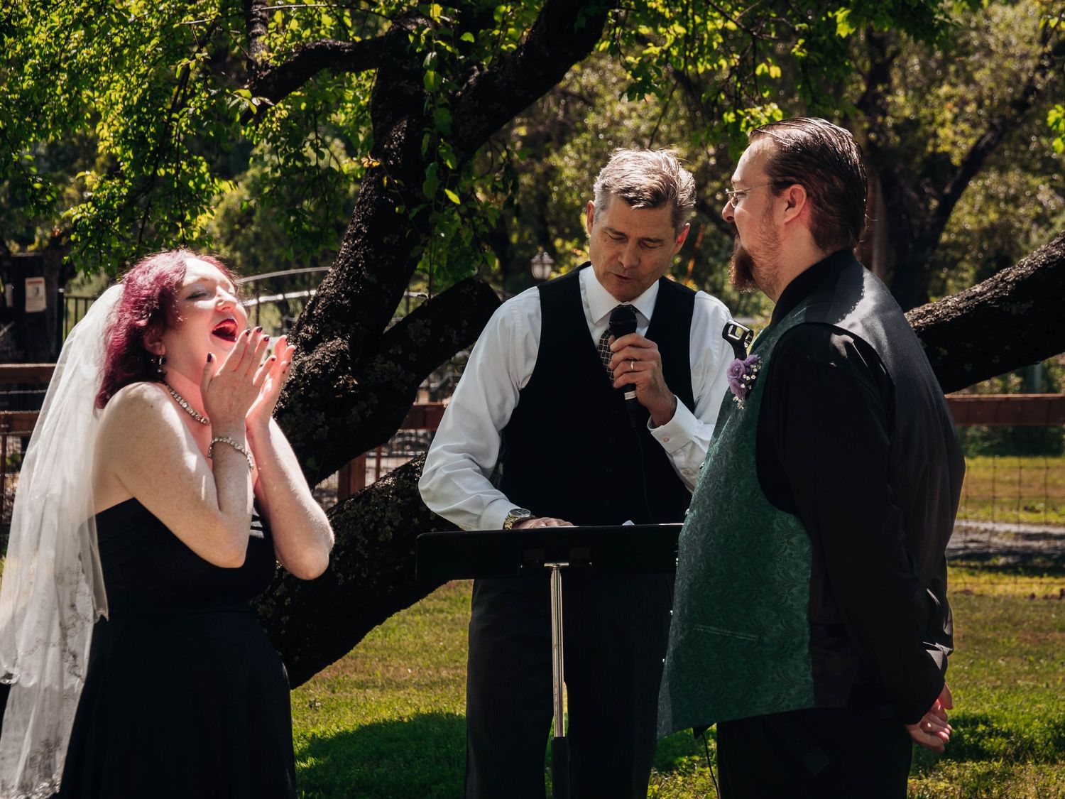Wedding ceremony participants gathered outdoors under trees during a sunny day.