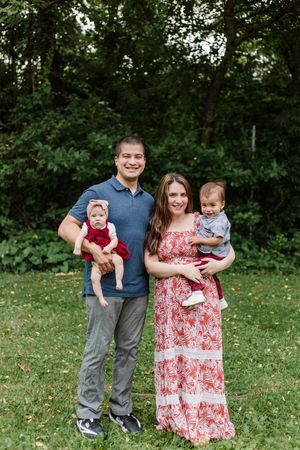 A family of four poses together outdoors on grass with trees in the background.