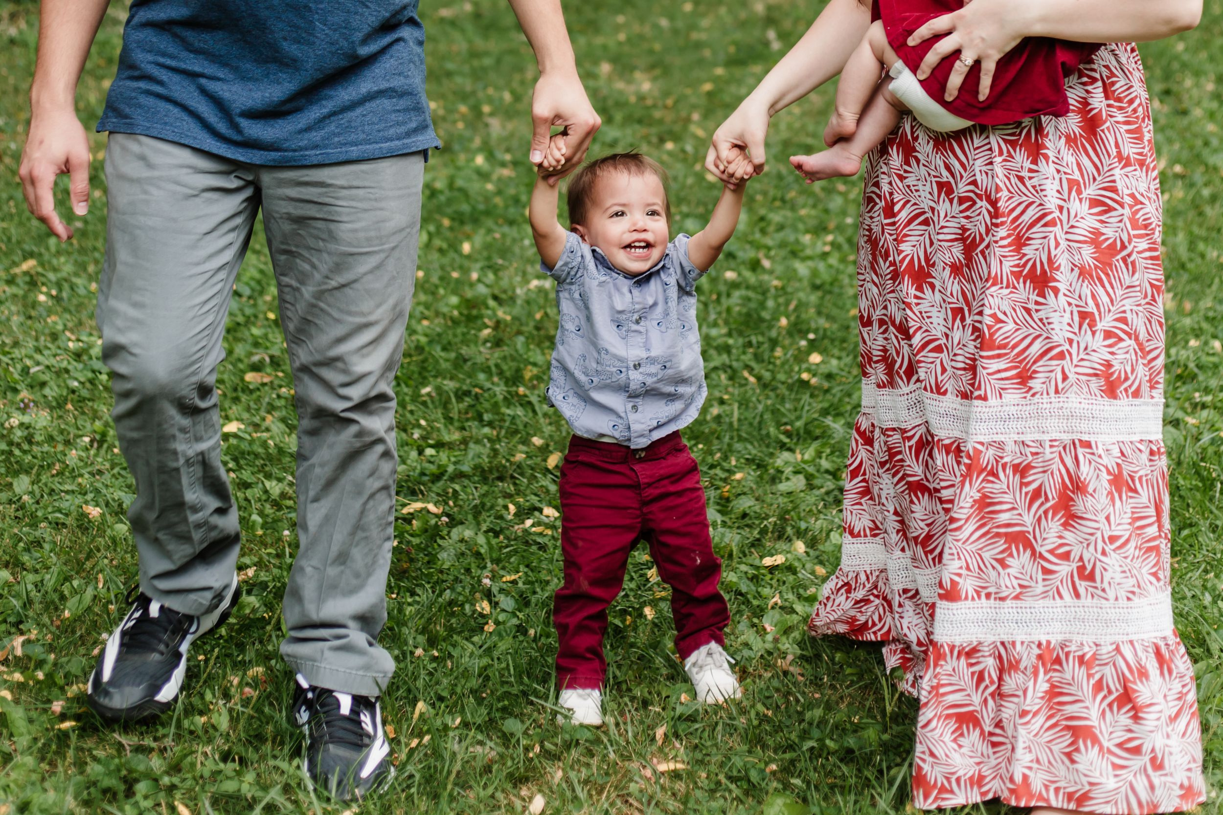 A small child in red pants jumps with joy while holding their parents' hands on a grassy lawn.