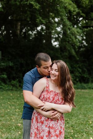 A couple embraces outdoors in a park setting, with lush green trees in the background.