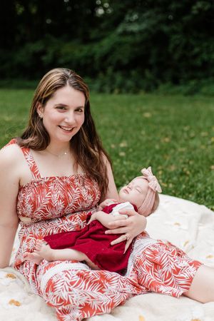 A woman in a red floral dress sits on a blanket outdoors holding a baby in red clothing.