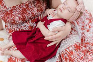 A sleeping baby in a red dress rests on white and red patterned fabric with delicate fern designs.