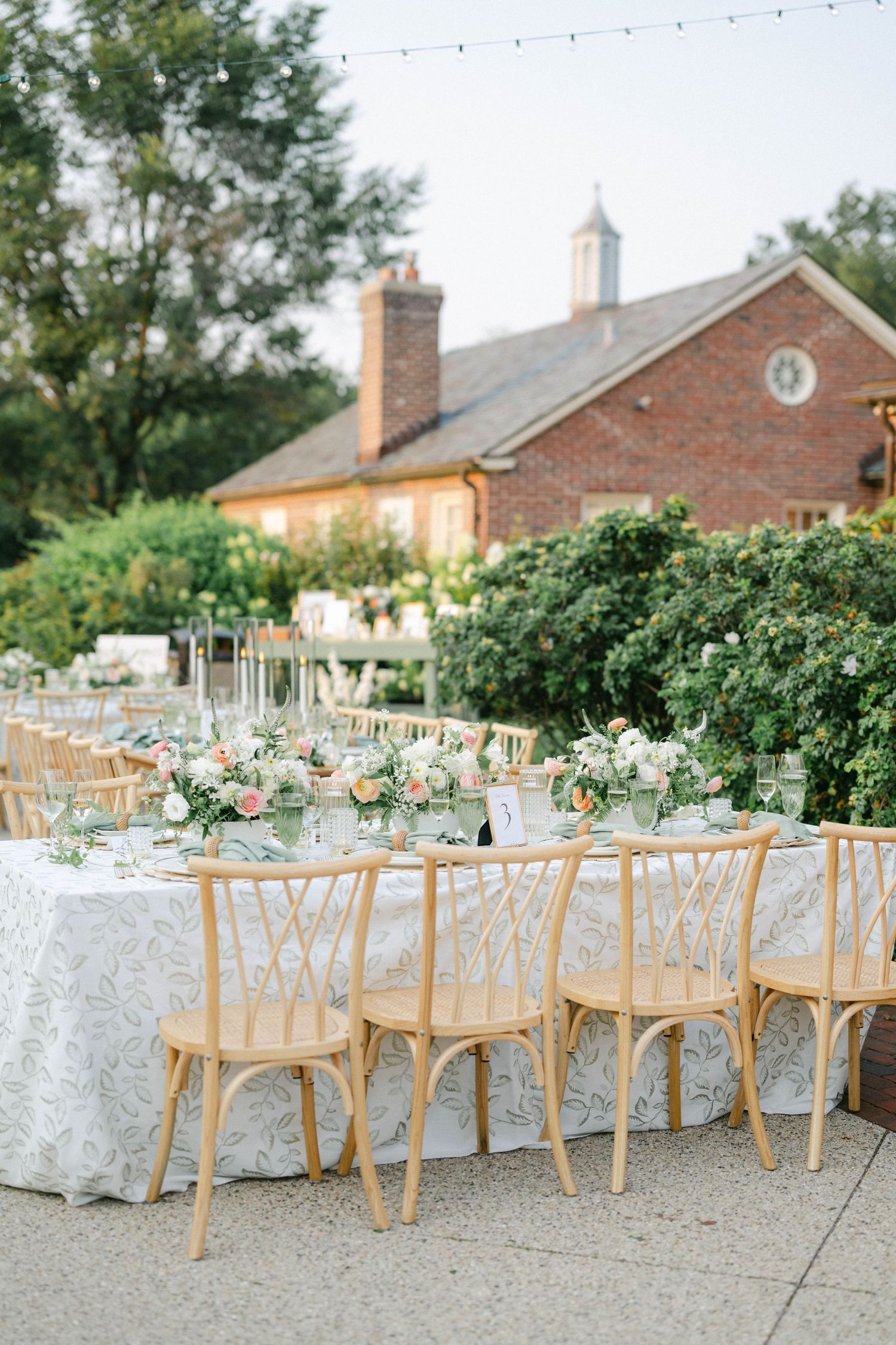Elegant outdoor wedding reception setup with wooden chairs and lace tablecloths against a brick building backdrop. - Elawa Farm