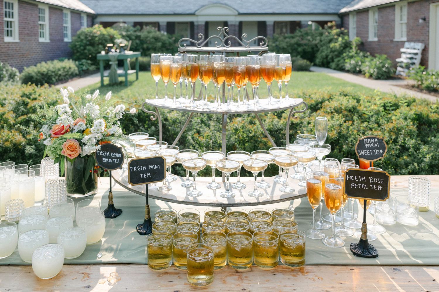 A rustic wooden table displays a food and drink spread in a garden setting with a brick building in the background. - Elawa Farm