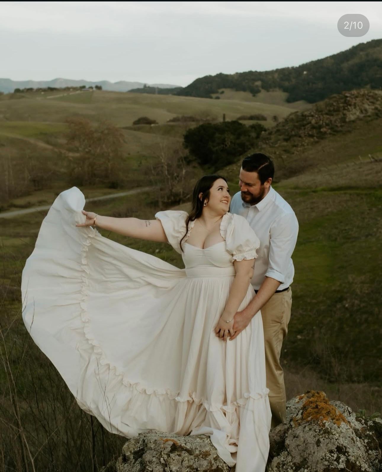 A couple poses against a scenic mountain landscape in romantic outdoor wedding attire.