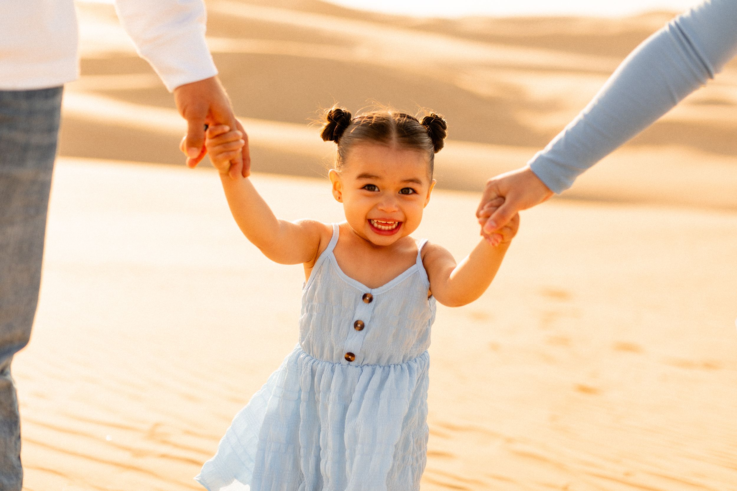 A toddler in a light blue dress holds hands with parents while walking in sand dunes.