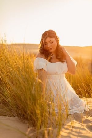 Woman in white dress poses gracefully among golden beach grass during sunset.