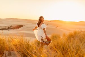 Woman in flowing white dress dances through sand dunes at golden hour.