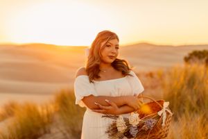 Woman in white dress holds basket while standing among beach grass at sunset.