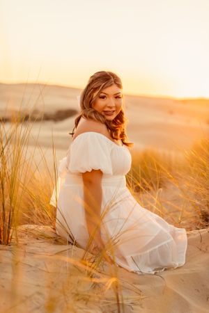 Woman in white dress sits among beach grass during golden sunset.