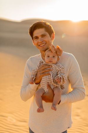 Parent holds infant wearing gingham outfit during desert sunset photo session.
