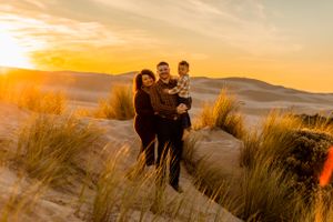 A family embraces while standing in tall grass during golden hour with dunes in background.