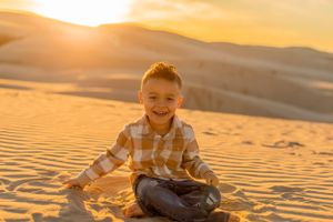 Young person sits cross-legged on sand dunes at sunset with a bright warm glow.