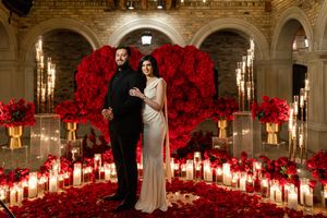Couple stands before giant red rose heart backdrop with romantic candlelit aisle in elegant stone archway setting.