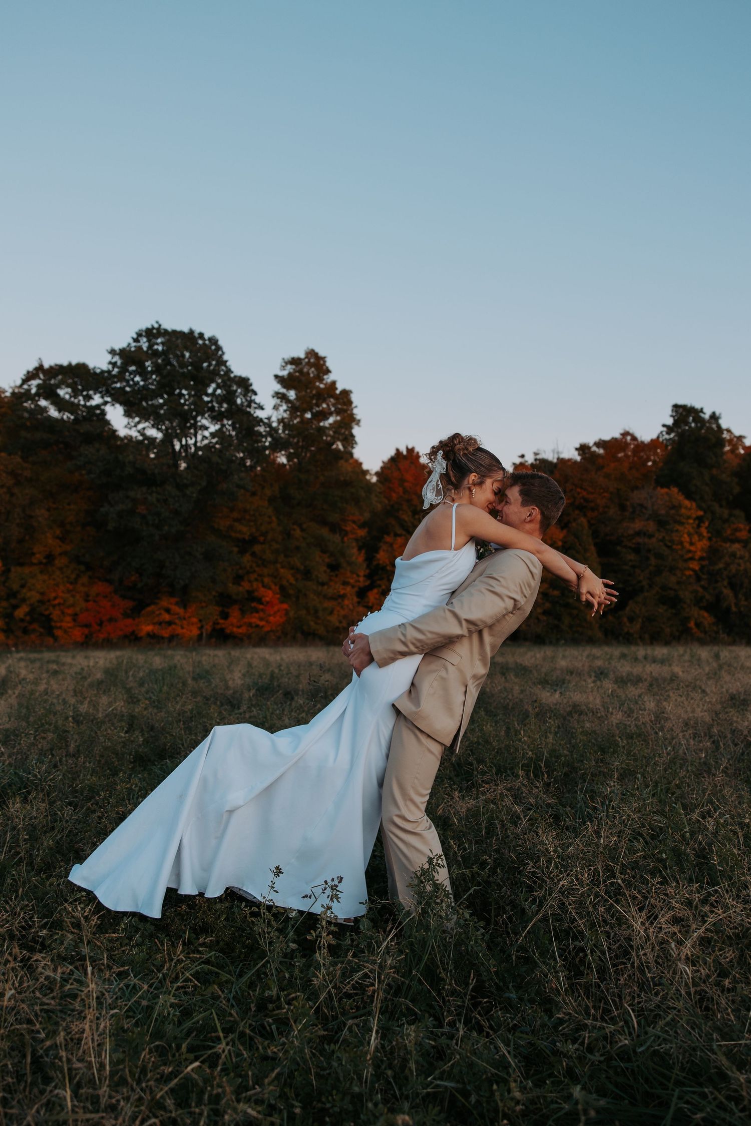 Couple in wedding attire dancing and dipping in a rustic field at sunset.