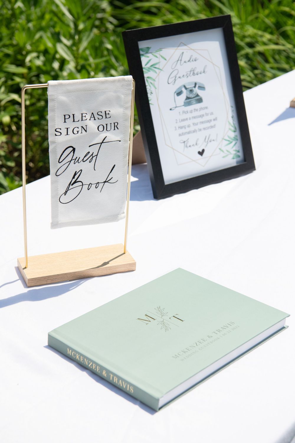 Wedding guest book and signage displayed on white tablecloth at outdoor venue.