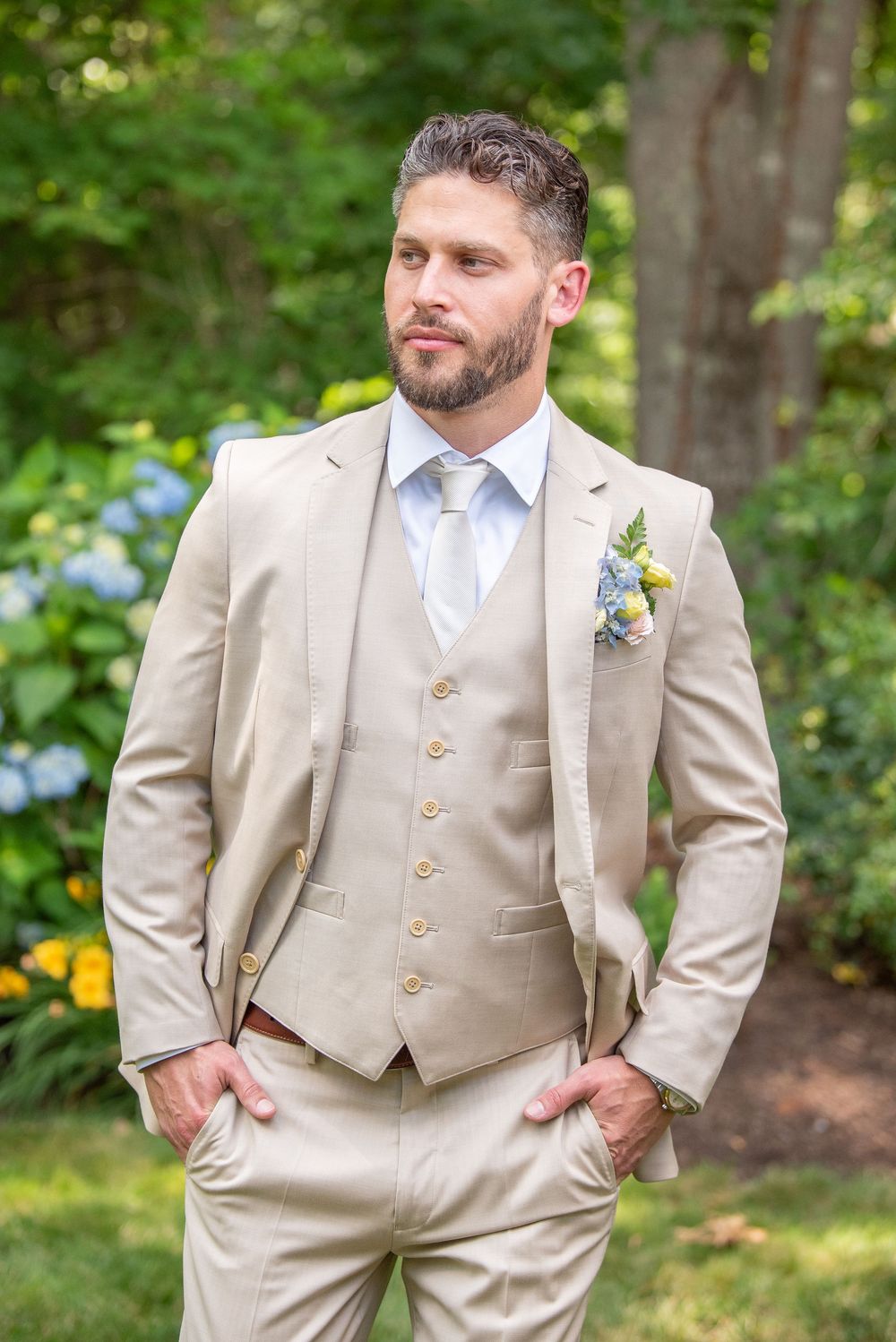 Groom poses in a three-piece beige suit with boutonnière against garden background.