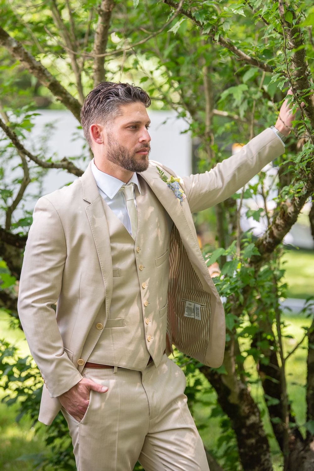 Groom in beige three-piece suit poses casually against trees in garden setting.