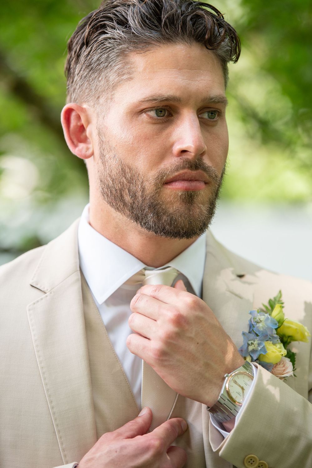 Side profile portrait of groom in beige suit adjusting tie outdoors.