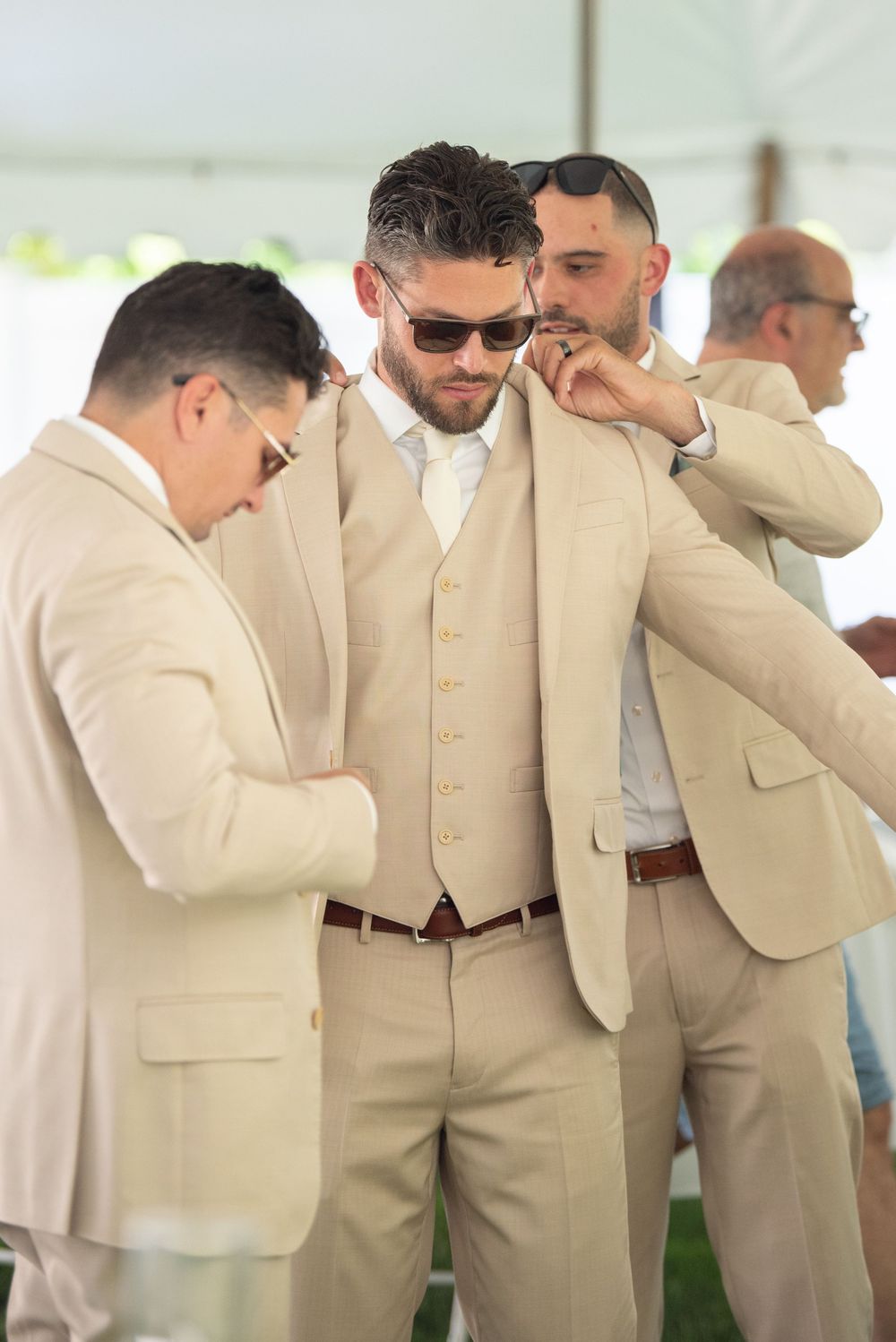 Groomsmen in cream colored suits stand together smelling their boutonnières in a fun wedding photo moment.