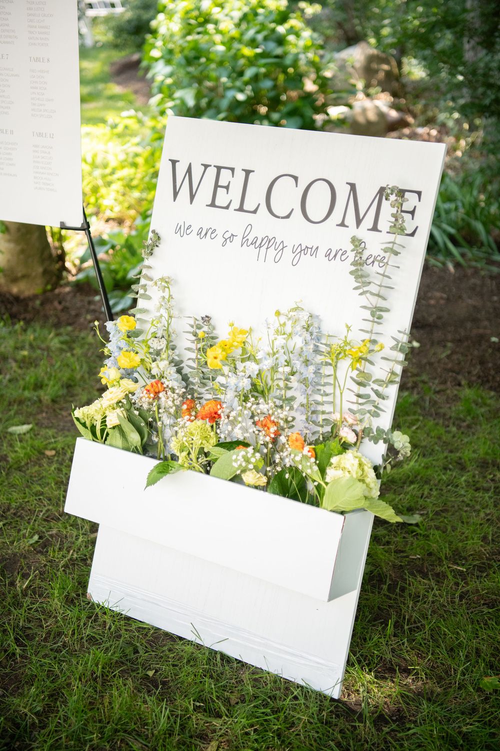 White welcome signs with floral arrangements displayed on grass for outdoor event.
