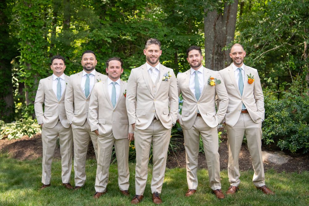 Wedding party in light beige suits poses together on grass with trees in background.