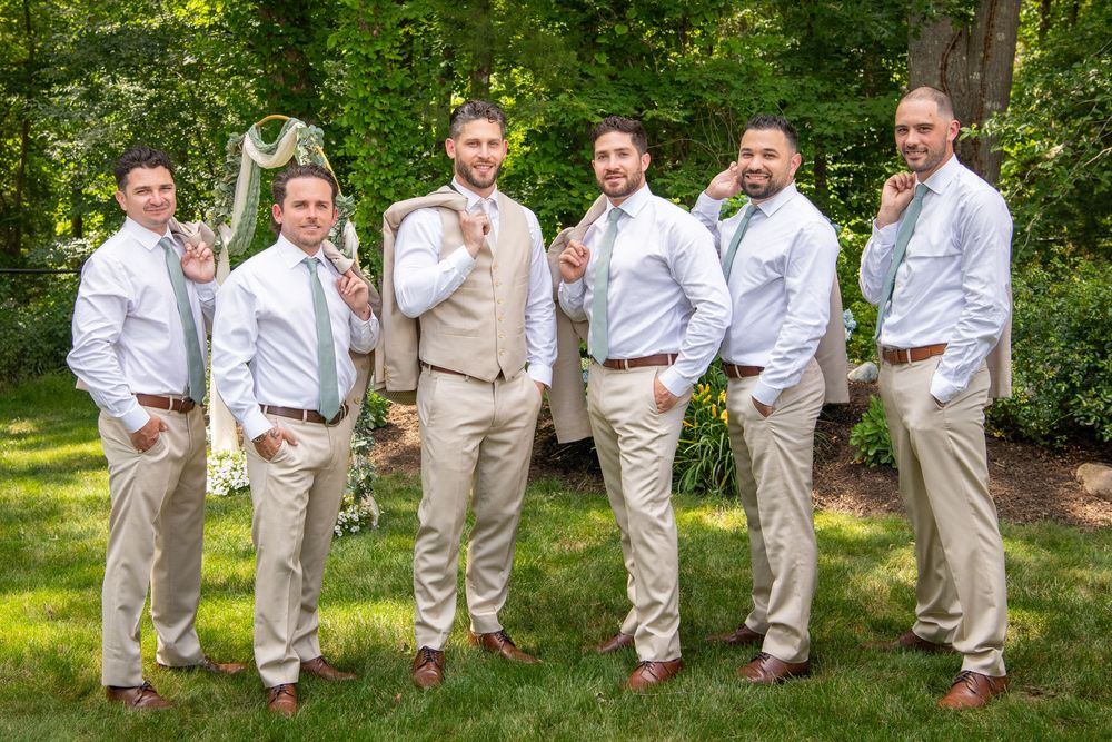 Groomsmen in tan suits pose playfully while jumping in unison on grass.