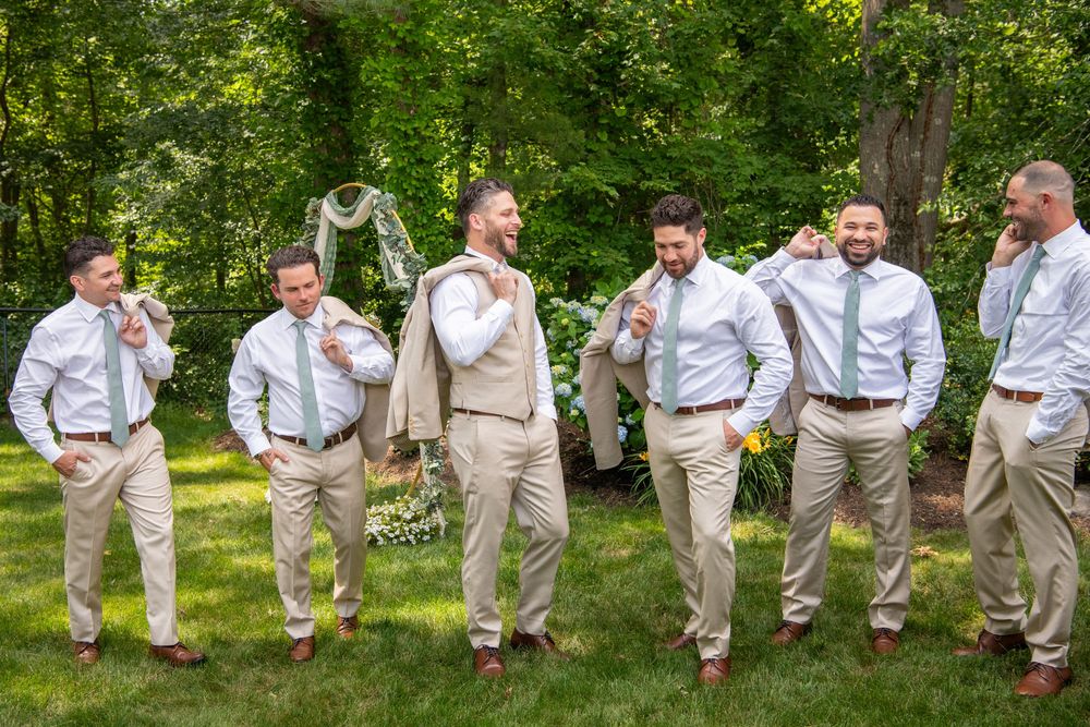Wedding party performs synchronized jumping poses outdoors in light colored suits.