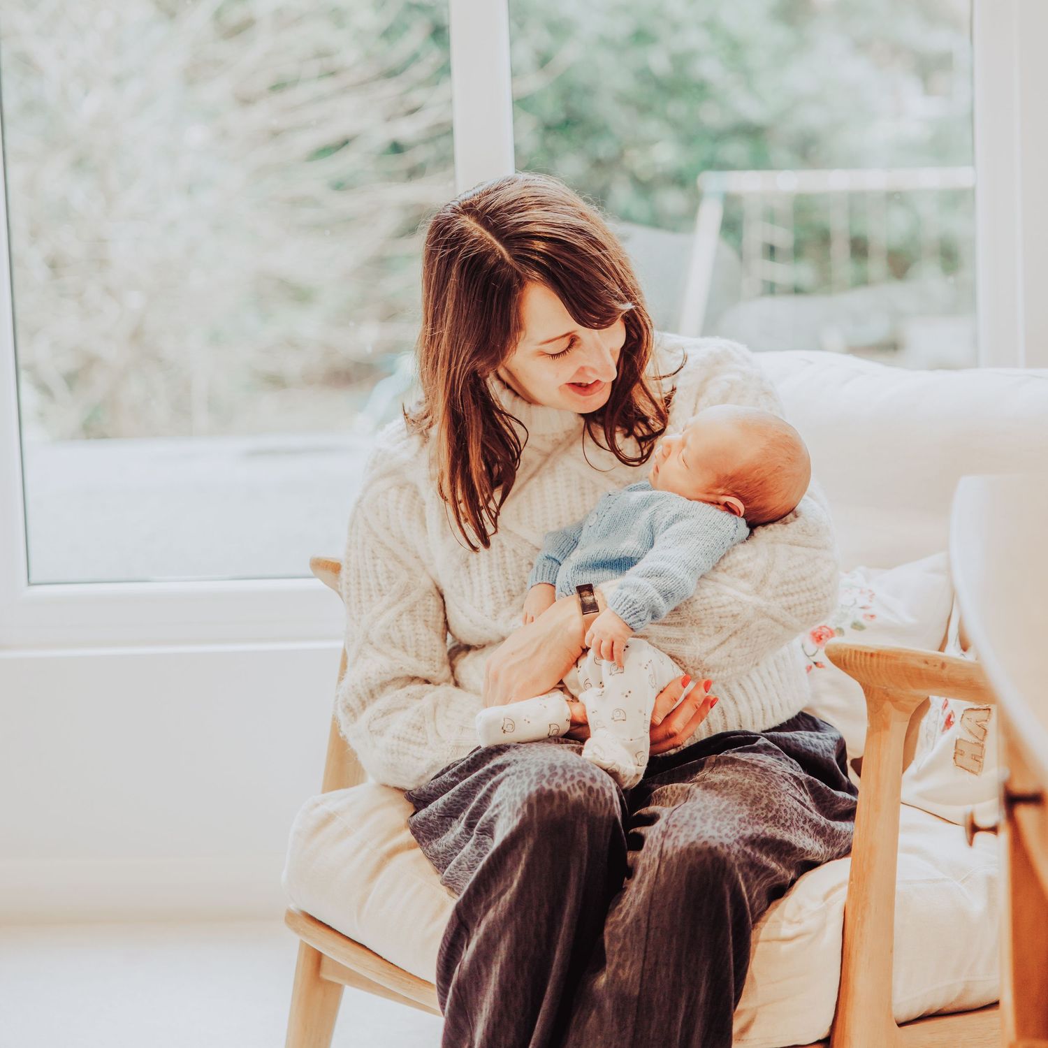 mum holding newborn baby on chair, looking lovingly at baby. window behind with natural light