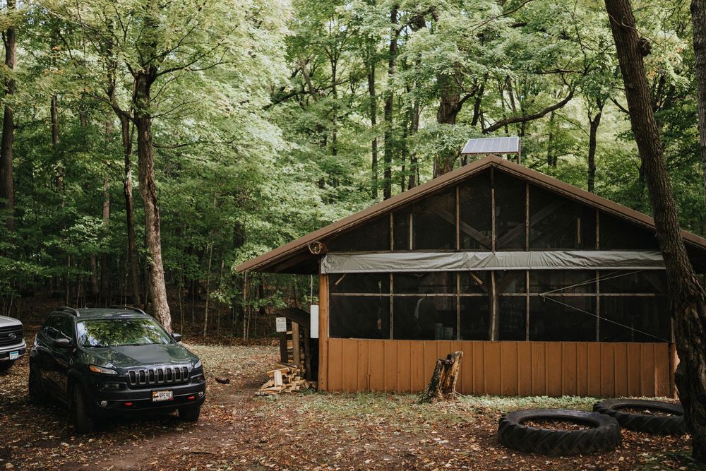 A rustic wooden cabin in the forest with a black Jeep parked outside surrounded by tall green trees.