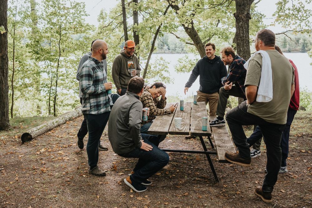 Group of people gathered around a campfire in a wooded area during outdoor social gathering.