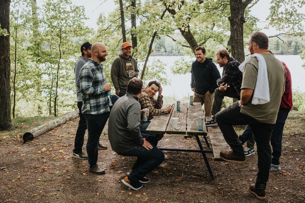 People gather in a wooded outdoor setting for a casual event with picnic tables and social interaction.