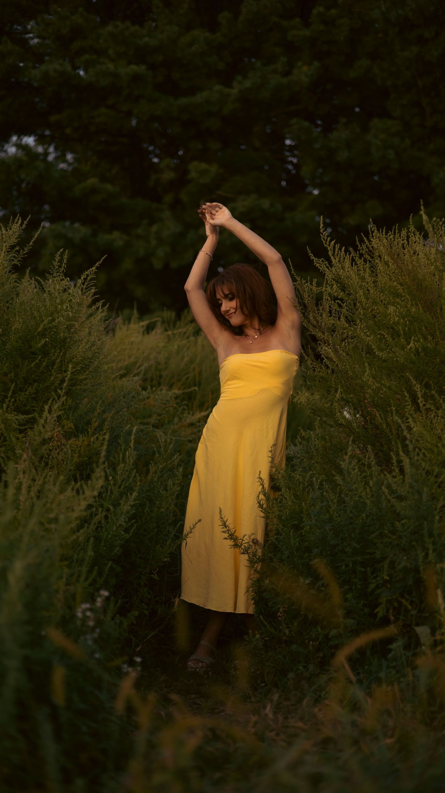 Figure in a yellow sundress stretches gracefully among tall grass at dusk with dark forest in background.