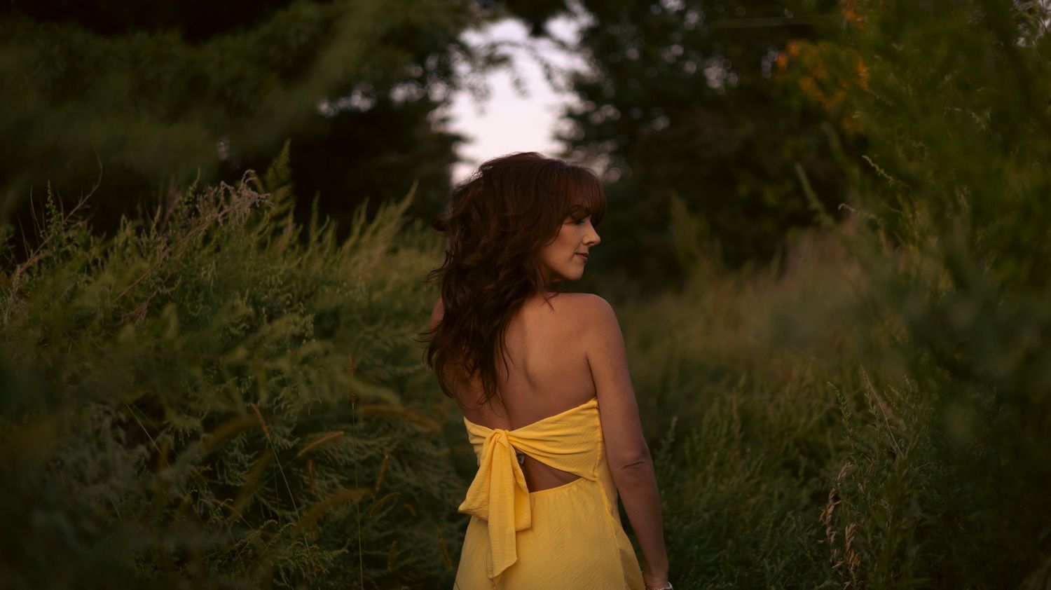 A person in a yellow dress walks through a lush green field at sunset creating a dreamy summer scene.