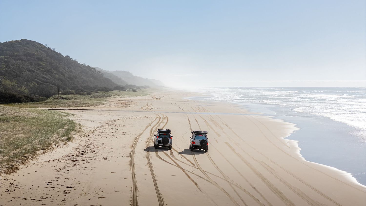 Aerial view of two 4WD vehicles driving along a remote sandy beach coastline.