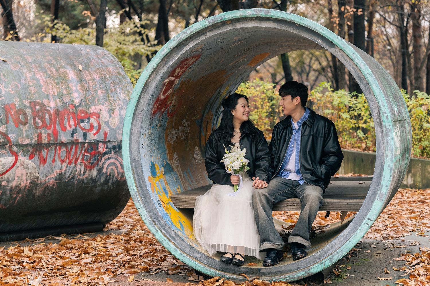 A couple sits together in a concrete tunnel surrounded by autumn leaves and graffiti covered walls.