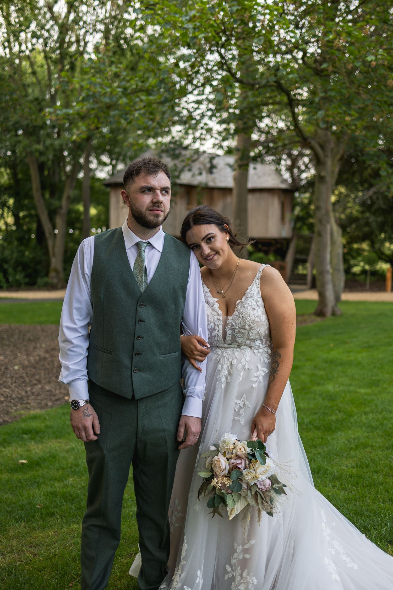 A romantic wedding couple poses together outdoors in a garden setting with lush green trees in the background.