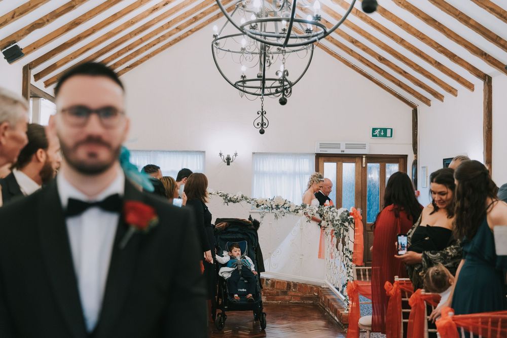 Wedding guests gather in an elegant venue with wooden beam ceiling and chandeliers during a ceremony.