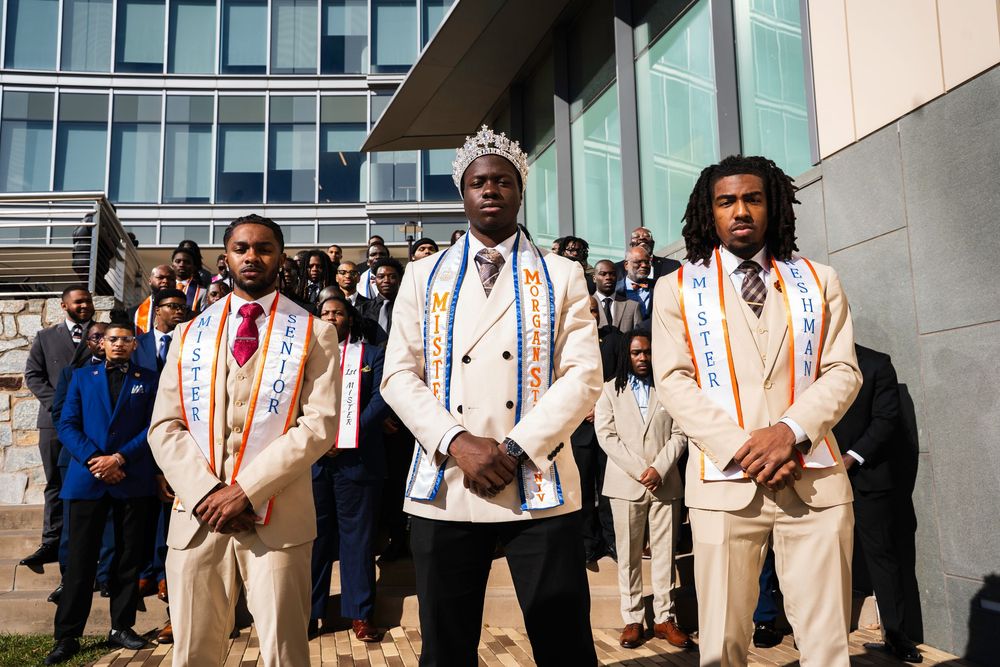 Group of people wearing beige suits and suspenders stand together in front of a modern glass building.