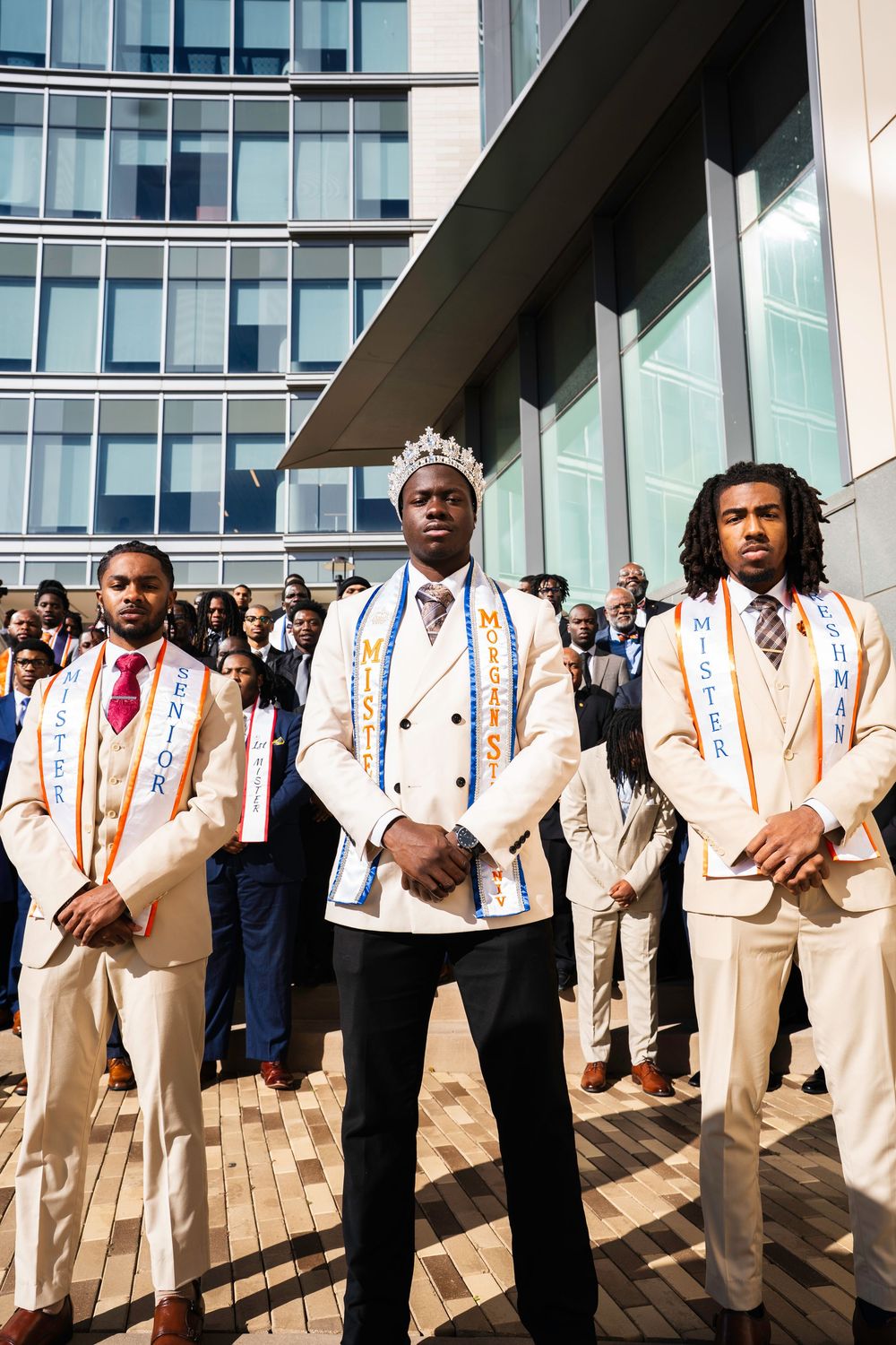A group of people in matching cream-colored suits with striped suspenders stand in a line outdoors near a glass building.