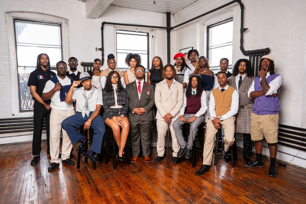 A group of people in formal attire pose together on a polished hardwood floor against a white wall.