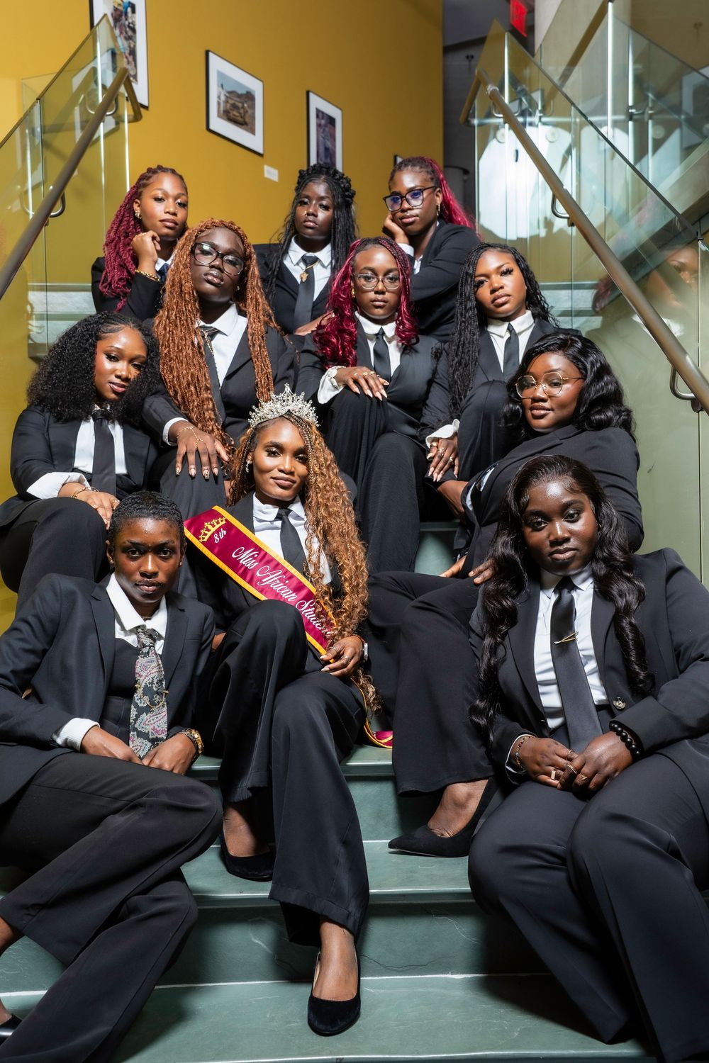 A group of people wearing black formal attire pose together on stairs in a stylish professional photo shoot.